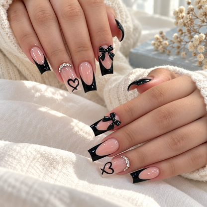 Black French tip press-on nails with 3D bows and hearts, worn on hands beside a cream knit throw